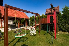 A wooden swing set with a tire swing and a slide, surrounded by a well-maintained lawn and a red-roofed building. - Two-story holiday homes with terraces, playground (Mielno)