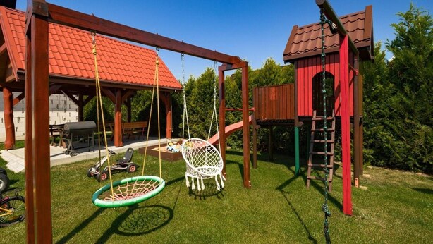 Un ensemble de jeux d’extérieur en bois, comprenant une balançoire avec un pneu et un toboggan, entouré d’une pelouse bien entretenue et d’un bâtiment au toit rouge.