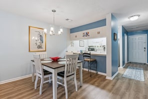 A dining area with a wooden table and chairs, a chandelier, and a kitchen in the background. - Brockinton Point Condo | Pool | Pets Allowed | Mid-Island Location | Full Kitchen | Patio | Deck (St. Simons Island)