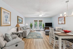 A living room with a sofa, a coffee table, a dining table with chairs, a TV, and a ceiling fan. - Brockinton Point Condo | Pool | Pets Allowed | Mid-Island Location | Full Kitchen | Patio | Deck (St. Simons Island)