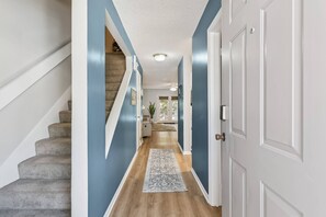 A hallway with a carpeted staircase, a door, and a view into a room with a plant. - Brockinton Point Condo | Pool | Pets Allowed | Mid-Island Location | Full Kitchen | Patio | Deck (St. Simons Island)