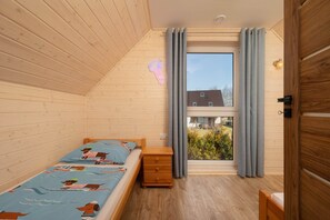 A bedroom with a wooden sloped ceiling, a bed with a blue ocean-themed bedspread, a wooden nightstand, a window with curtains, and a view of a house and greenery outside. - Year-round holiday home with fireplace and terrace (Mielno)