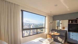 A hotel room with a large window, a bed, a dining area, and a kitchenette. - Studio 500m fr/ Vila Germanica, Blumenau Pin1908 (Blumenau)