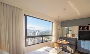 Quarto de hotel com uma janela grande, uma cama, área de refeições e uma cozinha compacta.