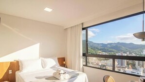 A hotel room with a bed, a nightstand, a lamp, a chair, and a large window with a city and mountains view. - Studio 500m fr/ Vila Germanica, Blumenau Pin1908 (Blumenau)