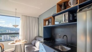 A modern kitchen with a built-in microwave, stove, and sink, a dining area with a table and chairs, and a large window offering a city view. - Studio 500m fr/ Vila Germanica, Blumenau Pin1908 (Blumenau)