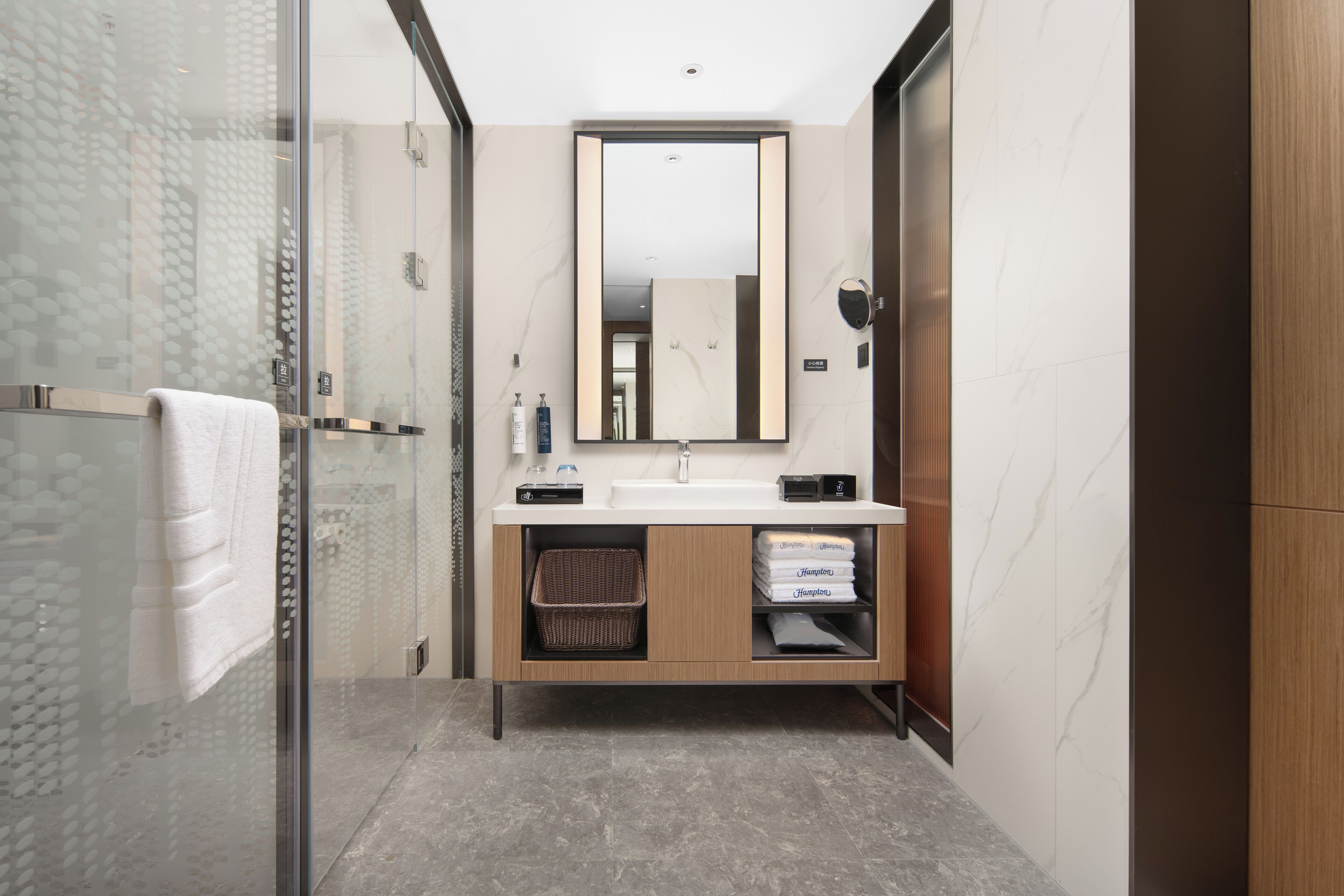 Un baño moderno con cabina de ducha de vidrio, tocador de madera con espejo y suelo de mármol.