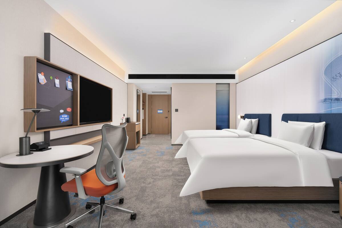Ein Hotelzimmer mit Bett, Schreibtisch mit Stuhl, Fernseher und einem an der Wand befestigten Bildschirm mit Informationen.