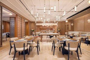 A spacious dining area with multiple tables and chairs, a central counter with a display, and pendant lighting. - Hampton by Hilton Jingdezhen Porcelain Center (Jingdezhen)