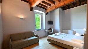Un dormitorio con cama, sofá, escritorio, silla, televisor y ventana con vista a follaje.