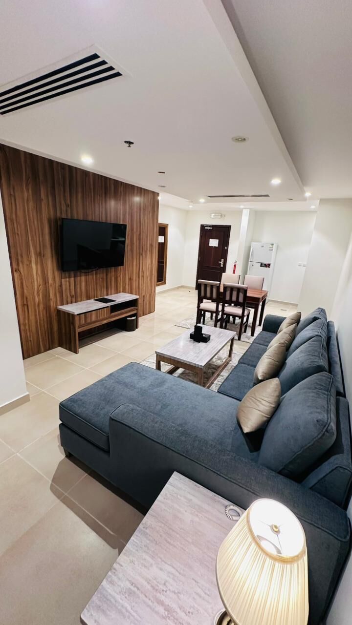 Ein modernes Wohnzimmer mit einer Eckcouch, einem an einer Holzwand montierten Fernseher, einem Essbereich mit Stühlen und einem Tisch und einer Küche im Hintergrund.