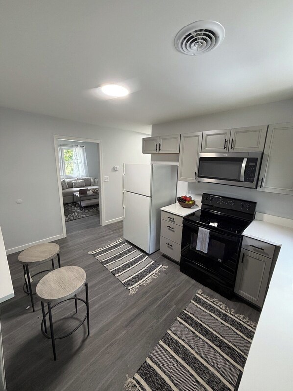 A compact kitchen with white cabinets, a black stove, and a microwave. There are two stools and a striped rug on the floor.