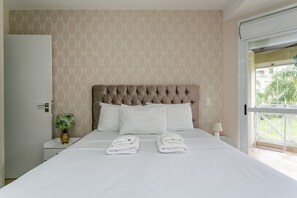 A neatly made bed with two folded towels, a nightstand with a lamp and a plant, and a window with a view of greenery. - WI-FI 350MB | Trapiche Lagoa da Conceição #LA12D (Florianópolis)