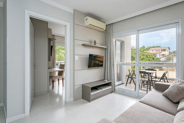 A modern living room with a flat-screen TV, built-in shelves, a sofa, and a sliding glass door leading to an outdoor patio. - Wi-Fi 500MB | 150m from the beach | Pool #IA37 (Florianópolis)