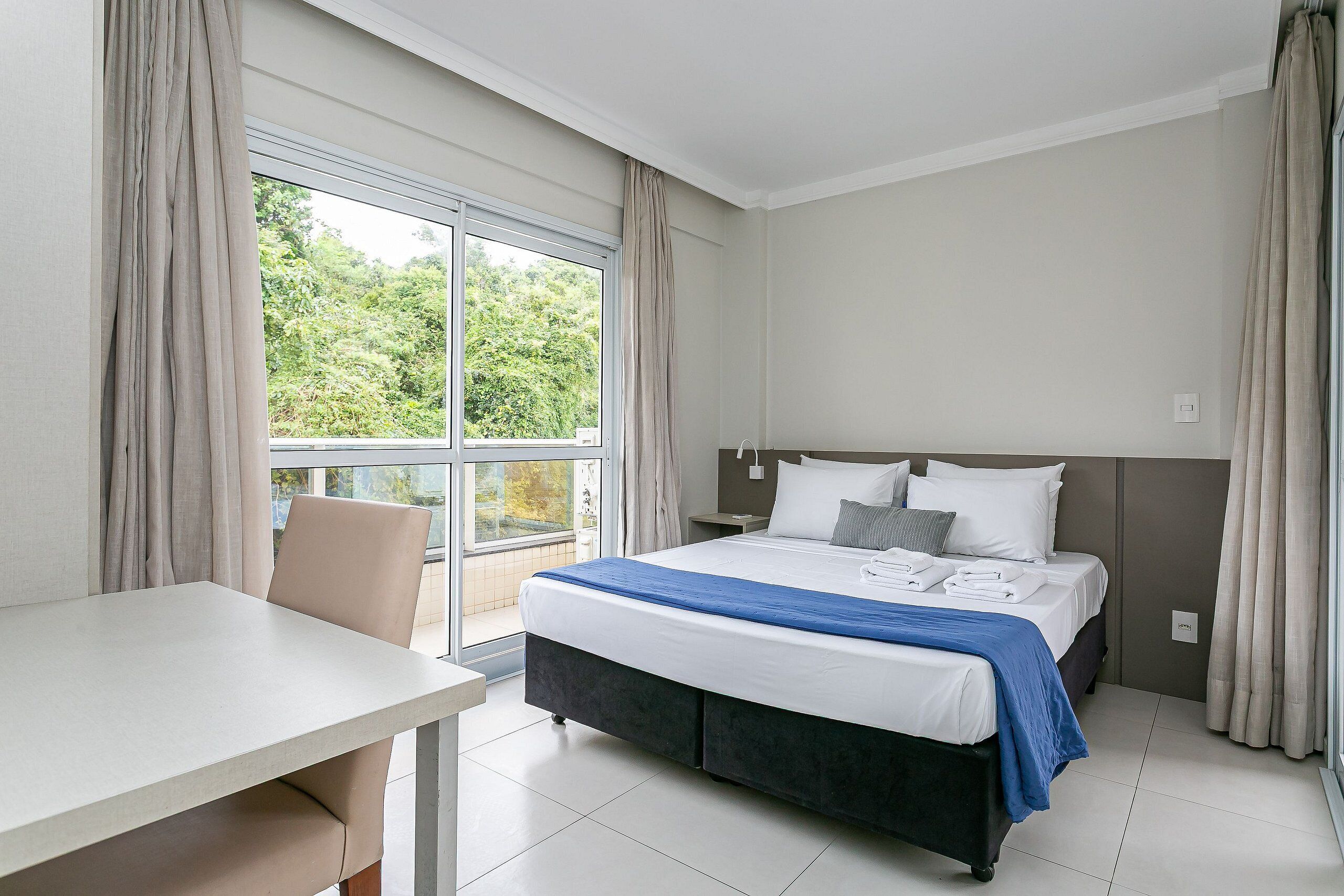 A hotel room with a bed, a table, and a view of greenery outside.