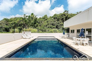 A rectangular swimming pool with clear blue water, surrounded by white lounge chairs and tables, set against a backdrop of lush greenery and a modern building. - Wi-Fi 500MB | 150m from the beach | Pool #IA37 (Florianópolis)