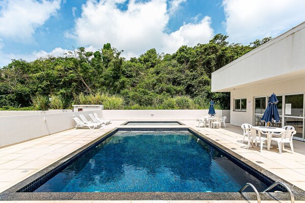 A rectangular swimming pool with clear blue water, surrounded by white lounge chairs and tables, set against a backdrop of lush greenery and a modern building. - Wi-Fi 500MB | 150m from the beach | Pool #IA37 (Florianópolis)