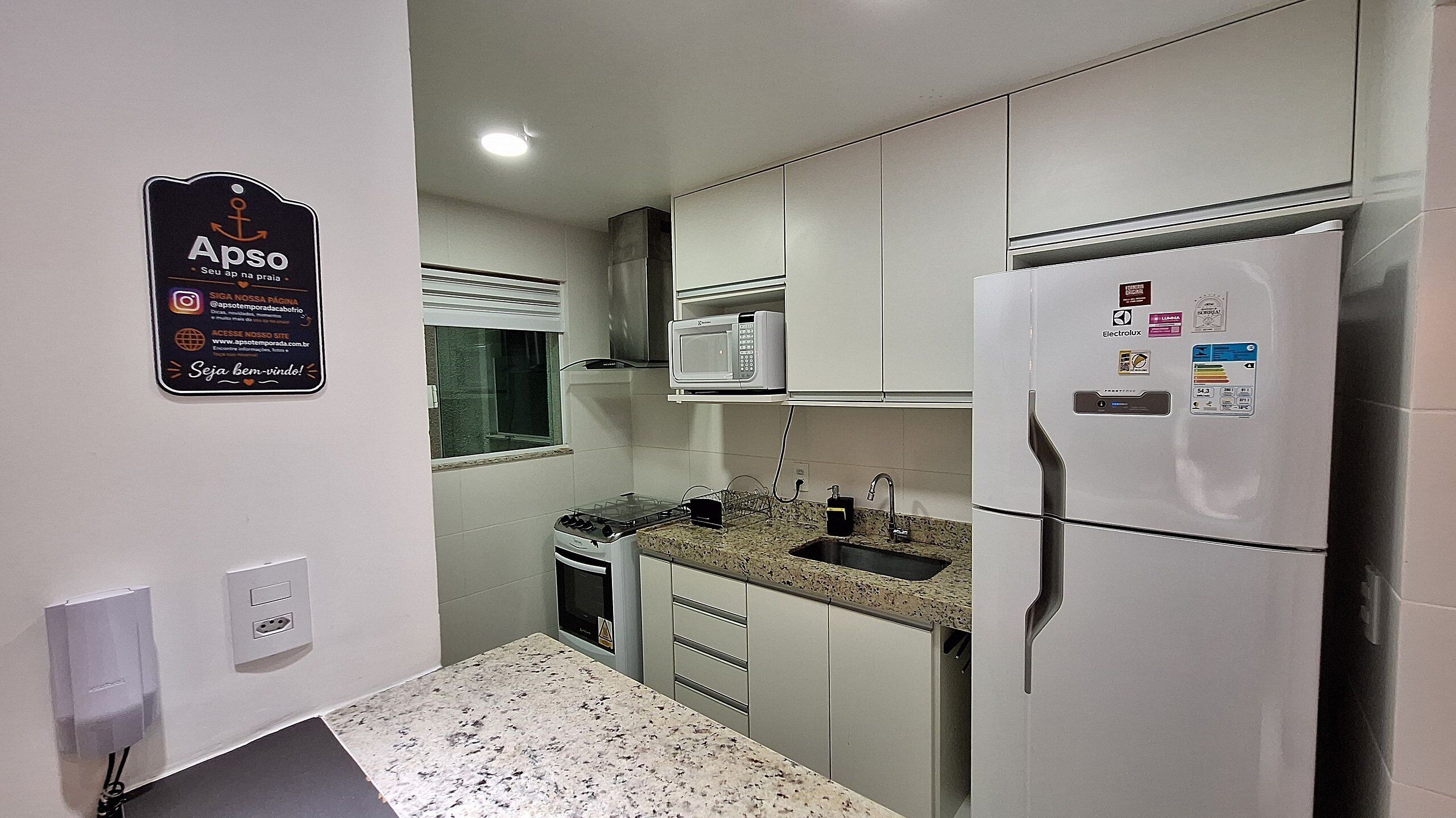 Una cocina compacta con armarios blancos, un microondas integrado, un refrigerador de acero inoxidable y una encimera de granito.