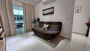 A modern living room with a brown sofa, a glass door leading to a balcony, a potted plant, and a framed picture on the wall. - O AP mais lindo da Praia do Forte 213 (Cabo Frio)