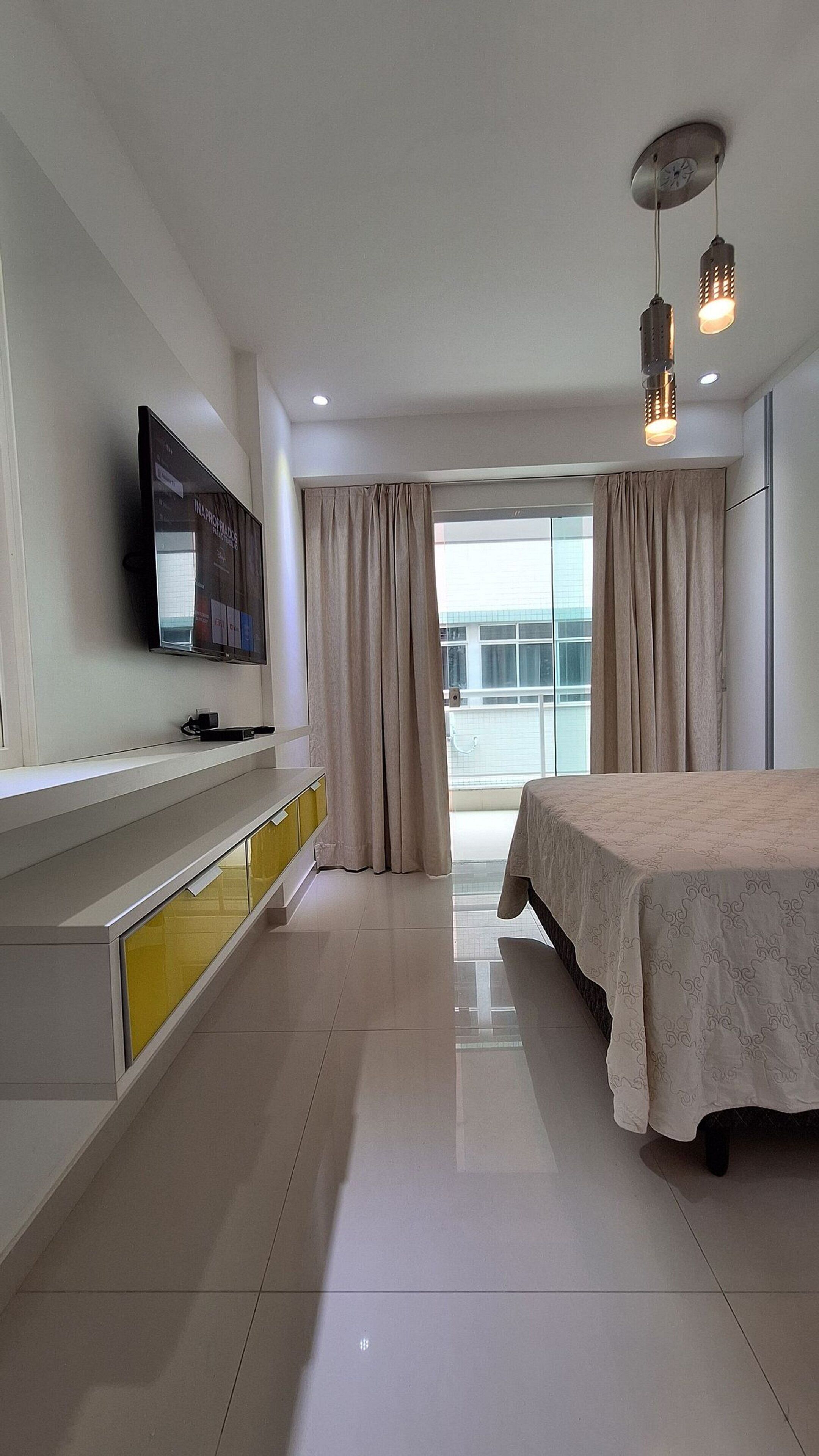 Quarto de hotel moderno com cama, televisão na parede, mesa de cabeceira e uma janela grande com cortinas.