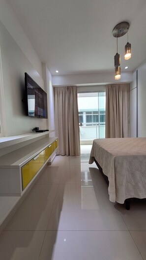A modern hotel room with a bed, a TV mounted on the wall, a bedside table, and a large window with curtains. - O AP mais lindo da Praia do Forte 213 (Cabo Frio)