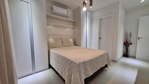 A bedroom with a bed, an air conditioning unit, a vase with flowers, and a door. - O AP mais lindo da Praia do Forte 213 (Cabo Frio)