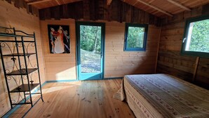 A wooden cabin room with a bed, a metal shelf, a painting, and two windows.