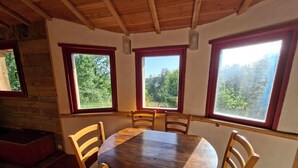 A wooden dining table with six chairs, large windows with red frames, and a view of greenery outside.