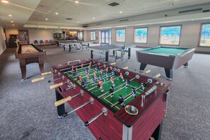 A pool table, foosball table, and ping pong table in a room with windows and a door.