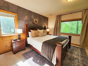 Un dormitorio con cama de madera, mesita de noche, cómoda, ventana con cortinas y vista a zonas verdes.
