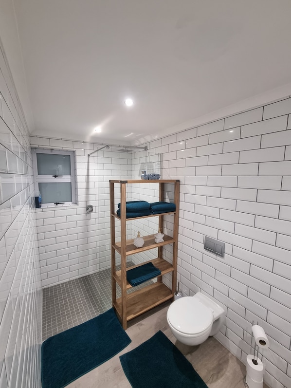 A bathroom with white tiled walls, a wooden shelf with towels, a toilet, and a shower area.