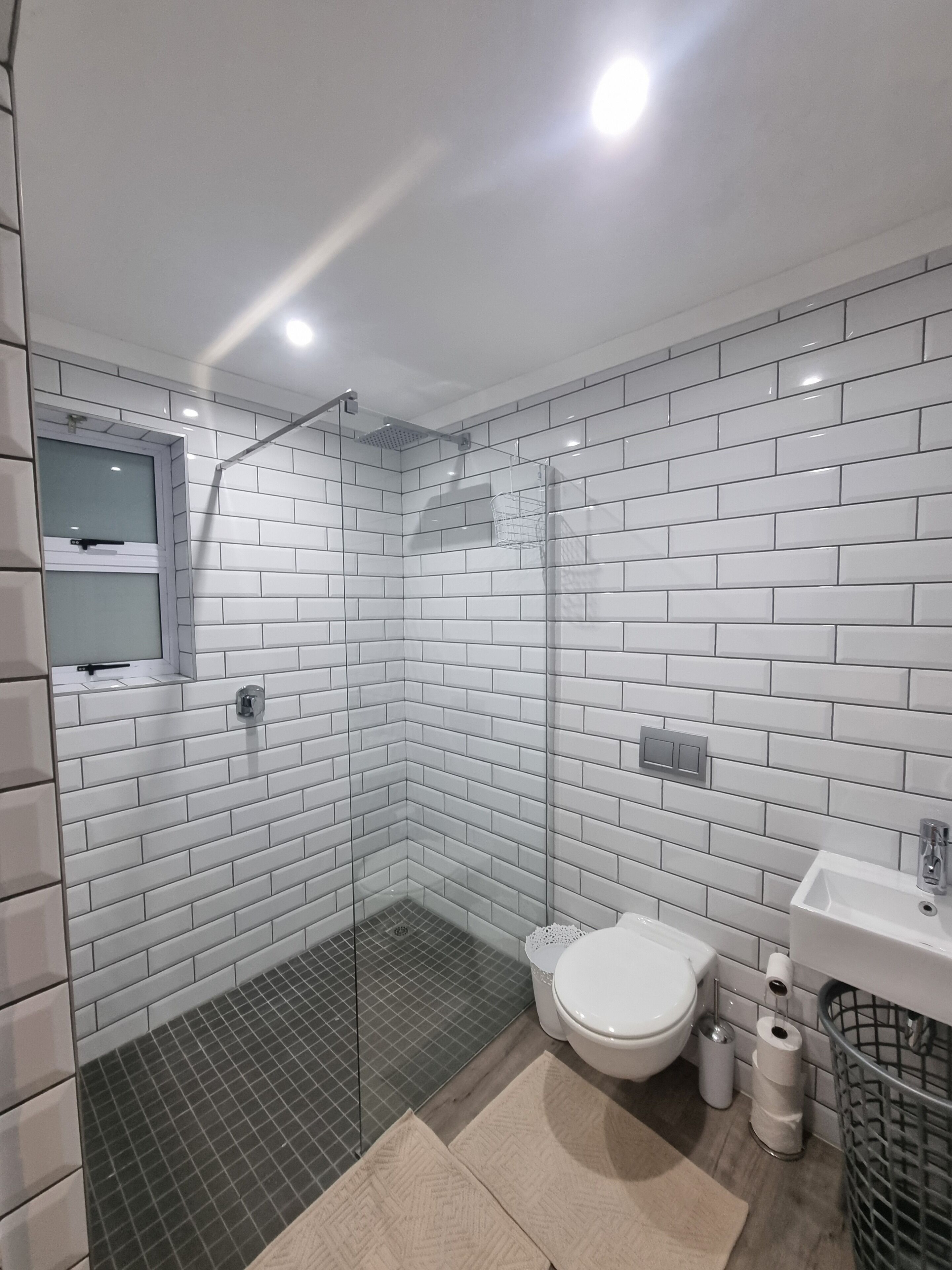 A modern bathroom with white subway tiles, a glass shower enclosure, a toilet, a sink, and a trash can.