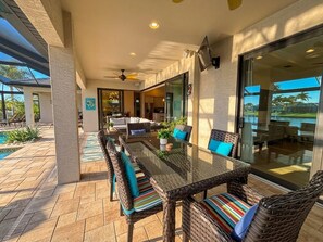 Un patio exterior con una mesa de vidrio y sillas de mimbre, un ventilador de techo y vistas a la piscina y la casa.