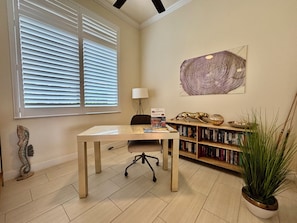 Ein Zimmer mit Schreibtisch, Stuhl, Bücherregal und einem großen Fenster mit Jalousien.