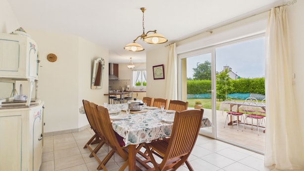 A dining area with a table set for six, wooden chairs, a kitchen in the background, and a sliding glass door leading to an outdoor patio. - Beach at 200m, family house for 7 with garden (Plouguerneau)