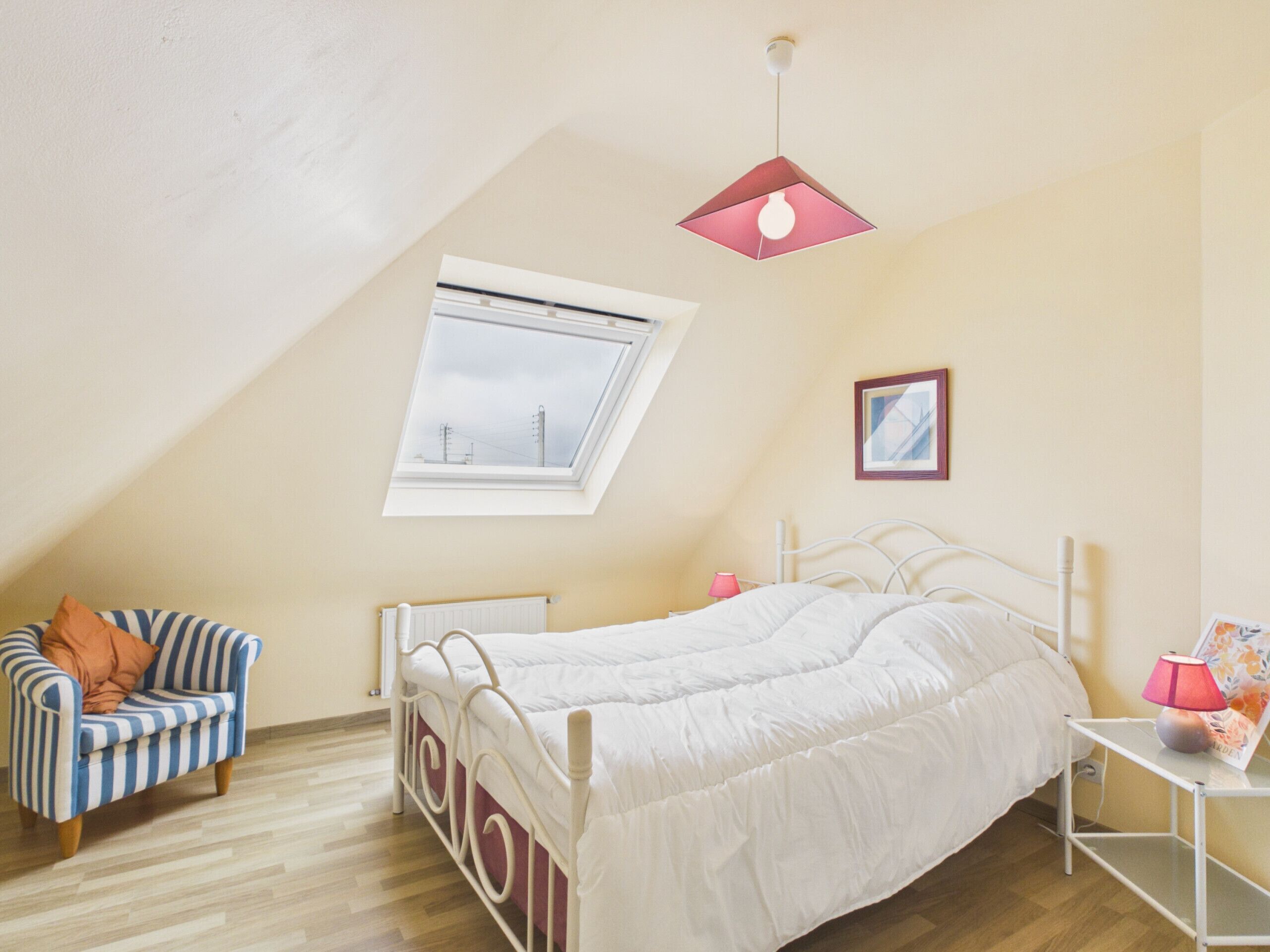 Une chambre avec un plafond en pente, un lit, une chaise, une table de chevet, une lampe, un tableau encadré et une fenêtre donnant sur des lignes électriques.