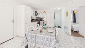 Een moderne keuken met witte kasten, een eethoek met een tafelkleed met patroon, en een zichtbaar bed in de aangrenzende kamer.
