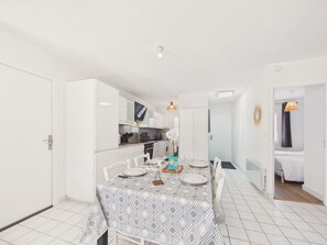 Eine moderne Küche mit weißen Schränken, ein Essbereich mit einem gemusterten Tischlein und ein sichtbares Bett im angrenzenden Zimmer.