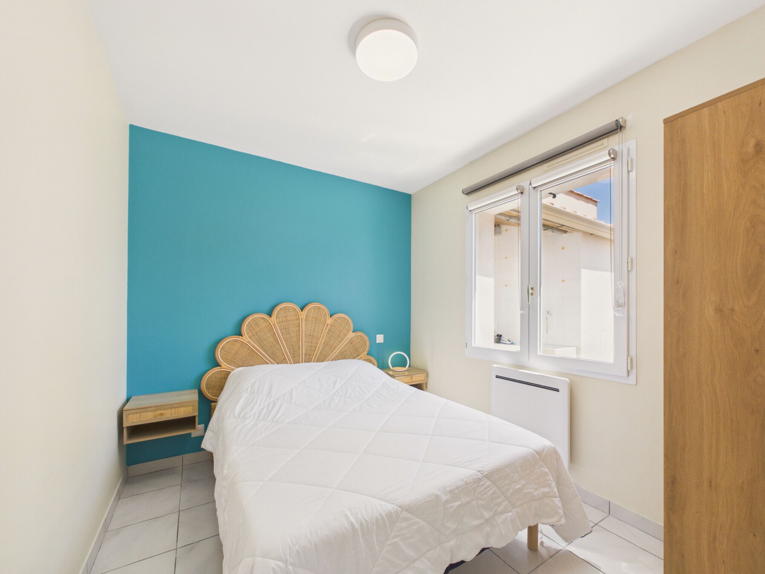 A bedroom with a bed, a window, a wooden wardrobe, and a small bedside table.
