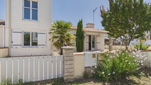 Une maison à deux étages, à l’extérieur blanc, avec une cour clôturée et un palmier.
