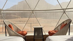 A room with two wicker chairs, a small table, and a view of a desert landscape through a large window. - Victoria Rum Luxury Camp (Wadi Rum)