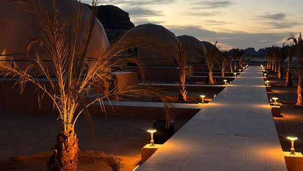 A pathway lined with palm trees and illuminated by ground lights, leading to a series of dome-shaped buildings. - Victoria Rum Luxury Camp (Wadi Rum)