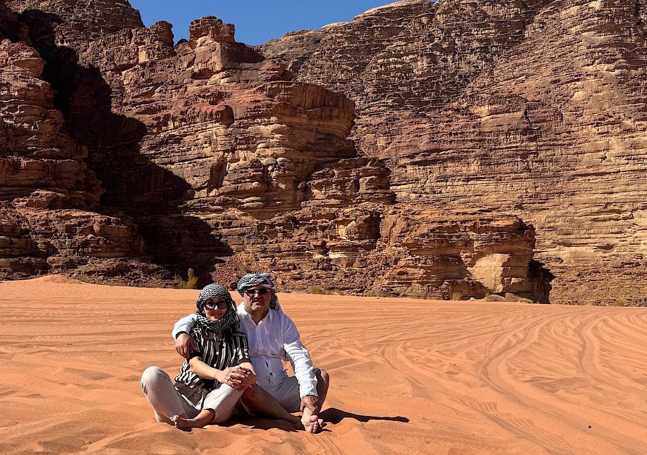 Two people sitting on a sandy surface with a rocky cliff in the background.