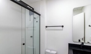 Uma casa de banho moderna com cabine de duche em vidro, sanita, lavatório preto e espelho.