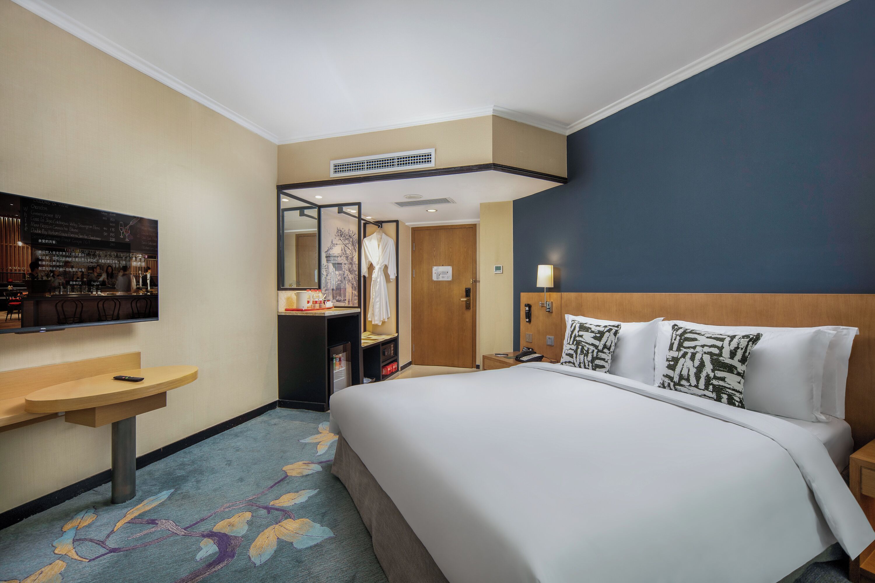 Habitación de hotel con cama, escritorio, silla, espejo, televisor montado en la pared y alfombra estampada con motivos florales.