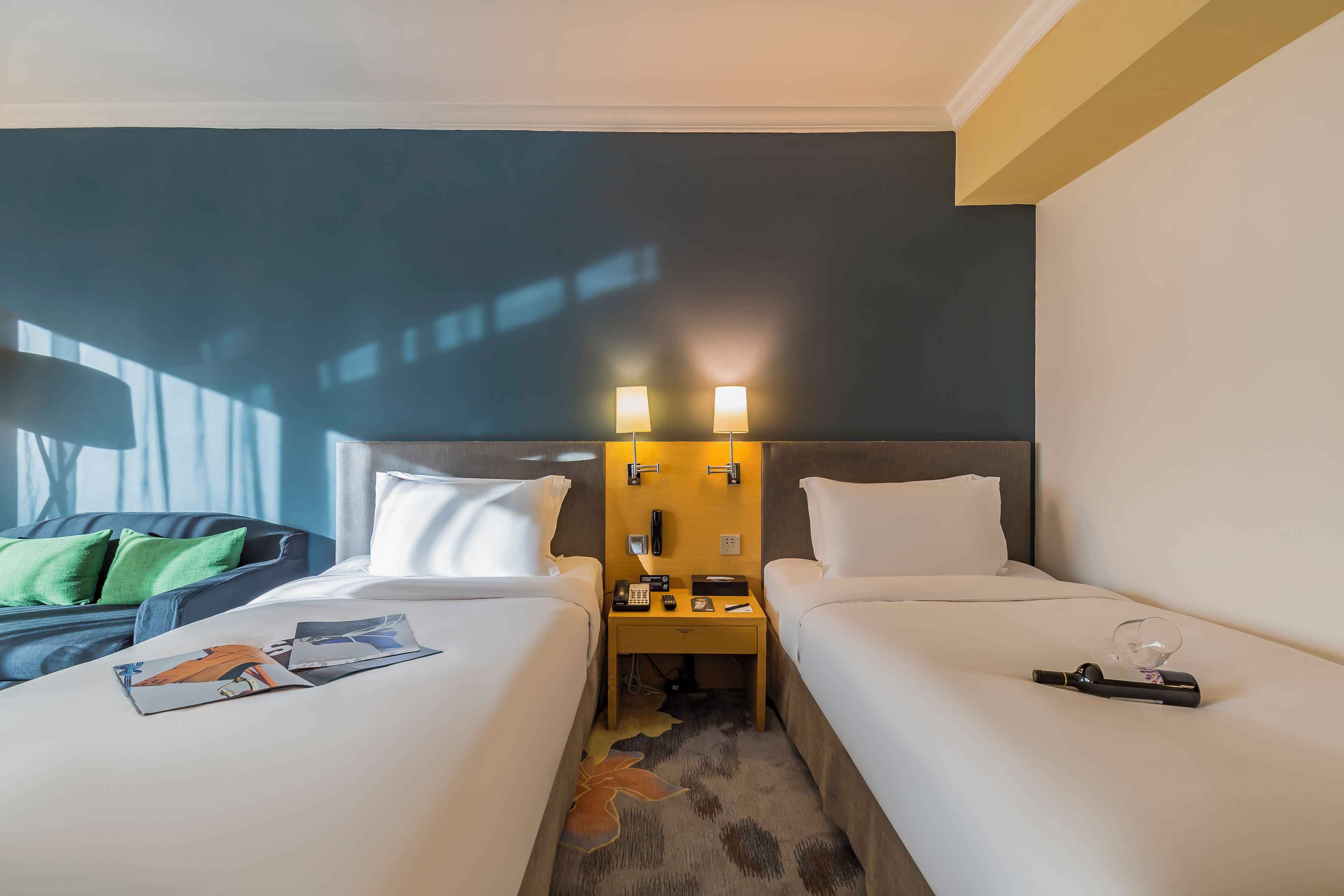 Habitación de hotel con dos camas, una mesita de noche con teléfono y lámpara, y una ventana con persianas.