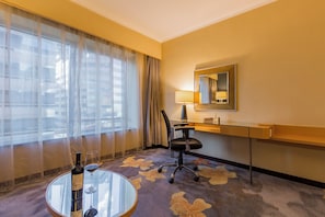Quarto de hotel com mesa de vidro, cadeira, escrivaninha, luminária, espelho e janela com cortinas translúcidas.