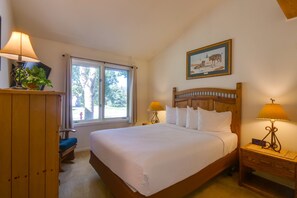 Un dormitorio con cama, dos lámparas de noche, una mesita de noche con un reloj, una ventana con cortinas y un cuadro en la pared.