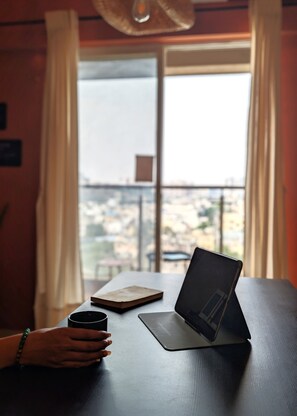 Ein Laptop auf einem Tisch mit einer Tasse und einem Buch, ein Fenster mit Vorhängen und Blick auf Gebäude.