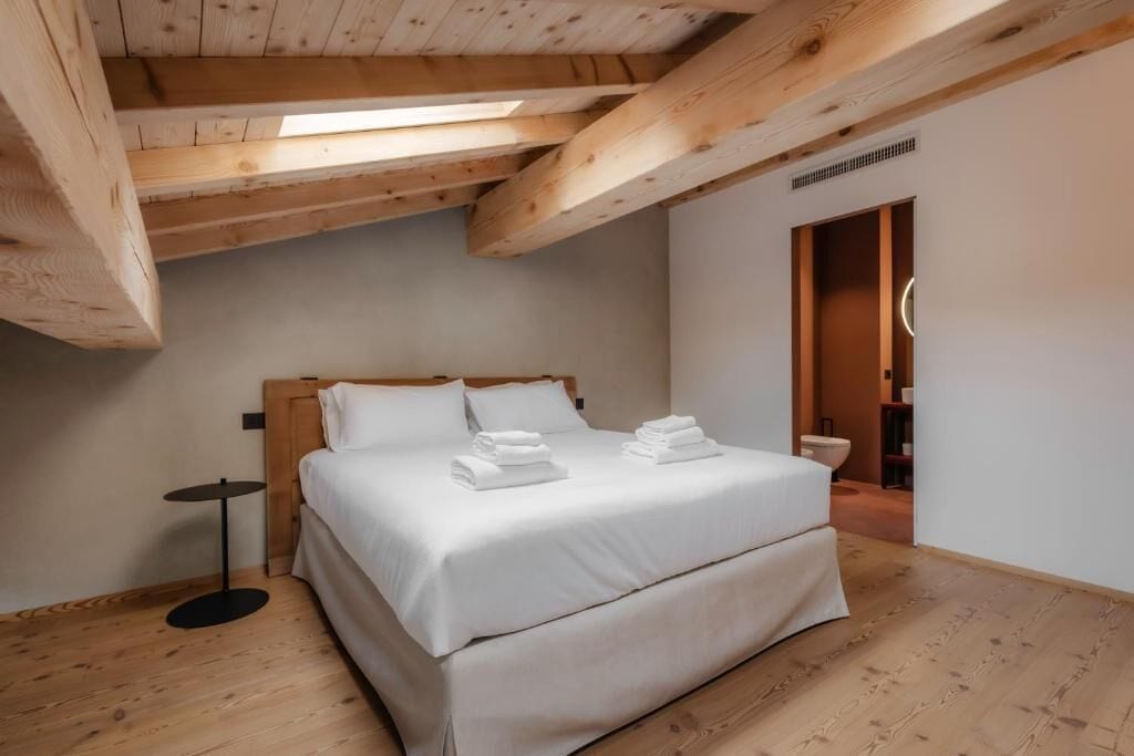 Una camera da letto con soffitto in legno, un letto con lenzuola bianche, un piccolo tavolino rotondo e una porta che dà su un bagno.